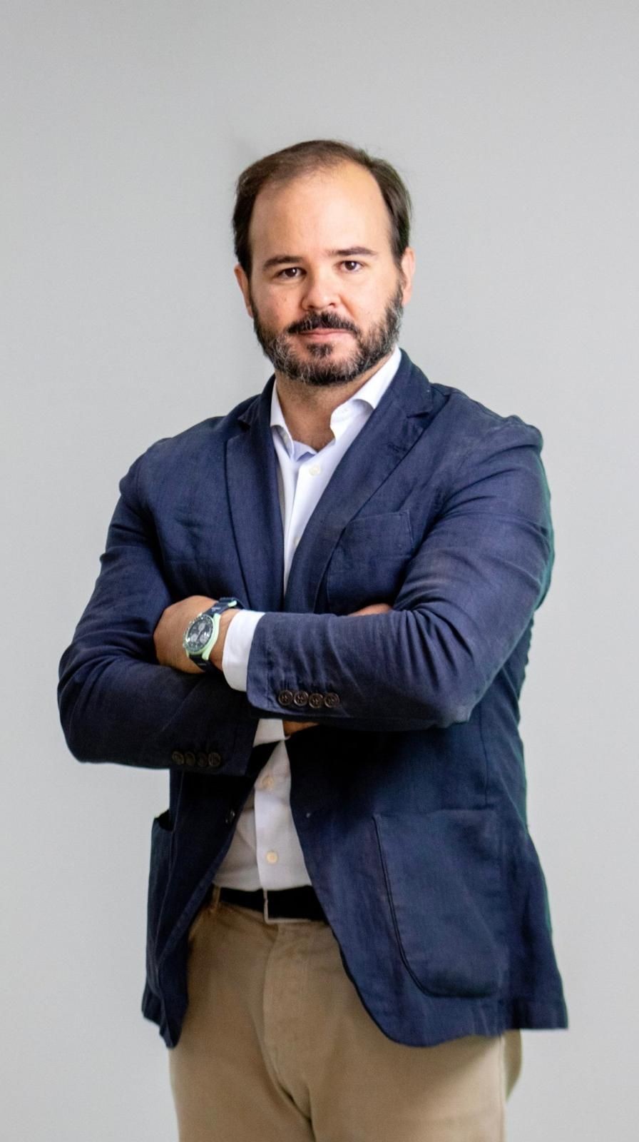 Man in a navy blazer with crossed arms standing against a gray background.